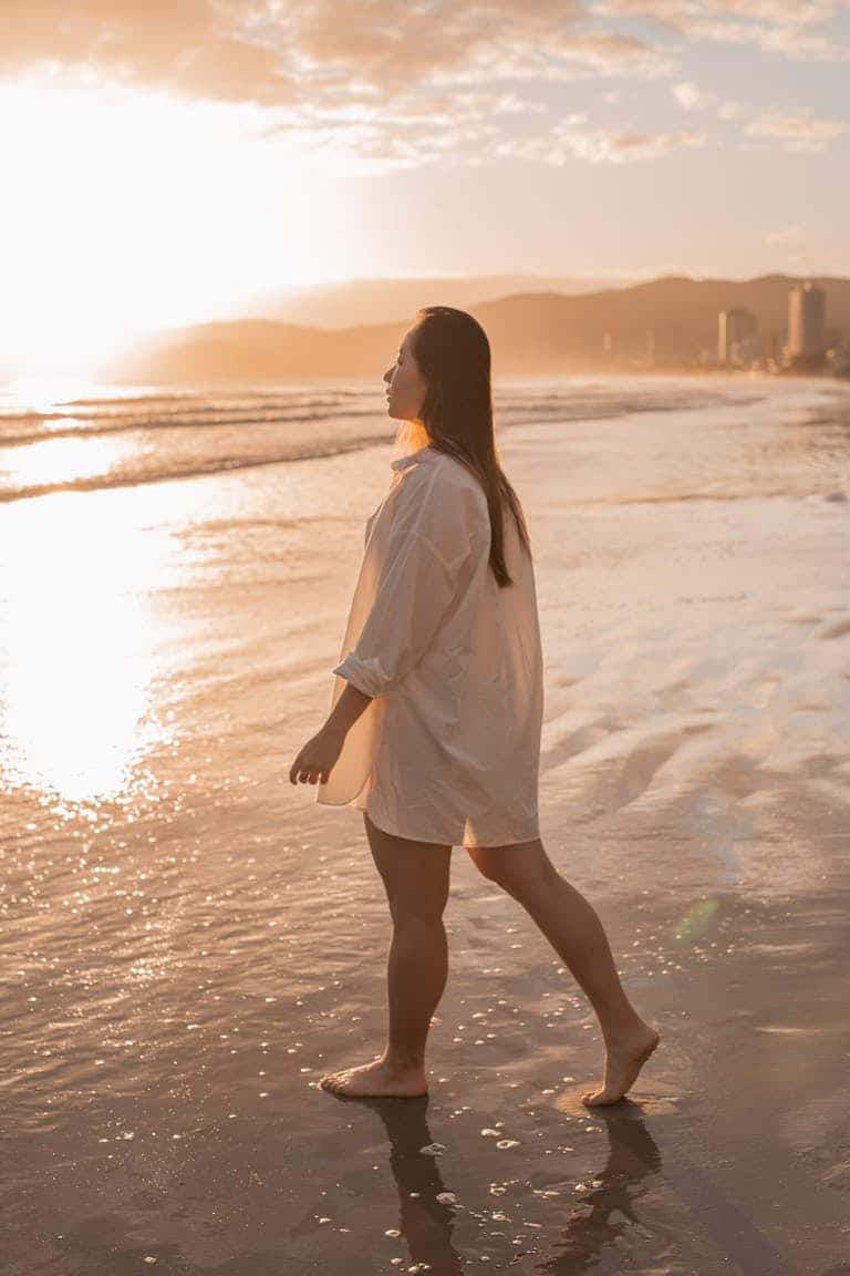 Woman walking barefoot on the beach at sunset, enjoying a tranquil summer evening.